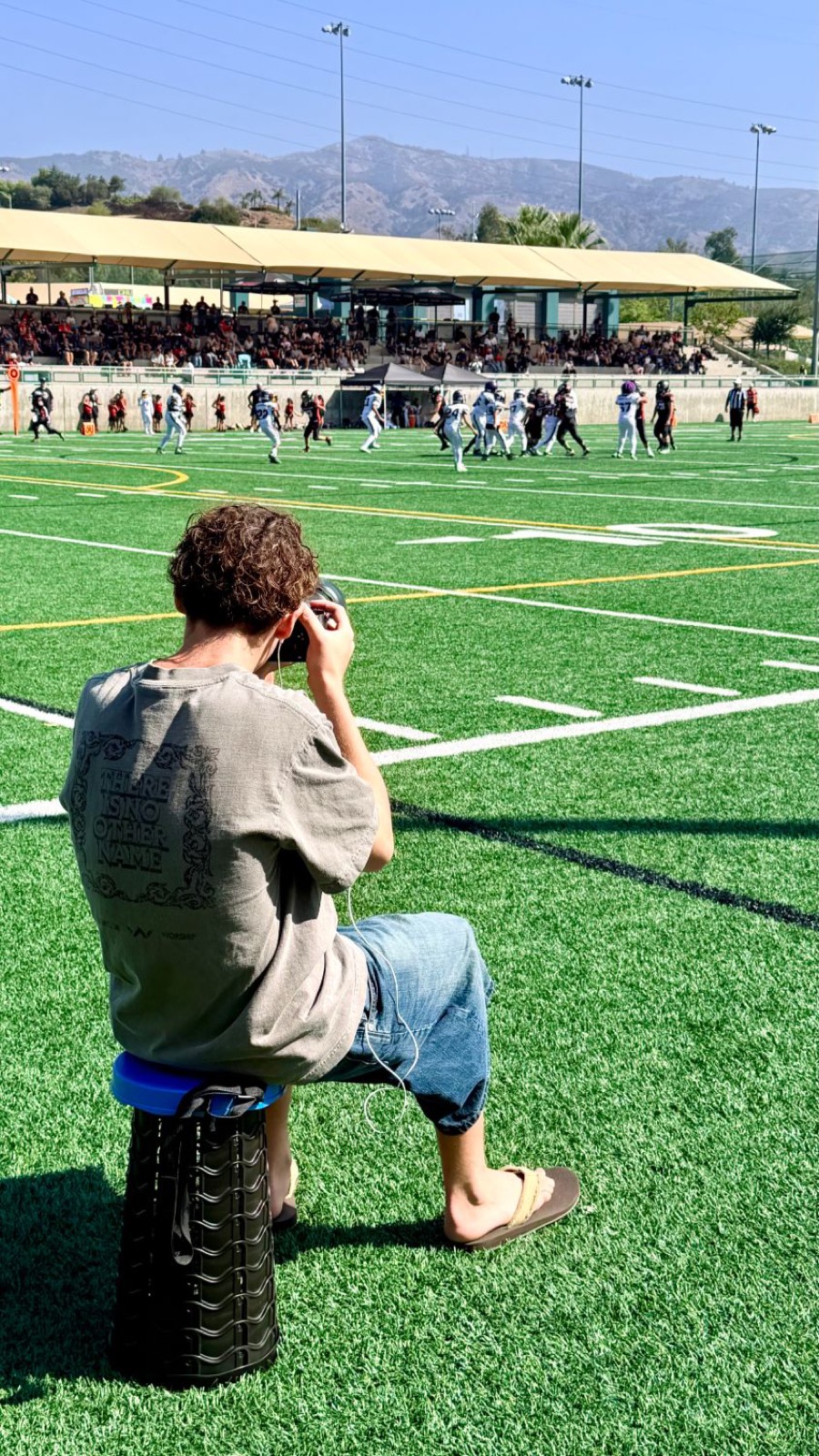 Hudson sitting on a stool at the edge of the field, photographing a football game